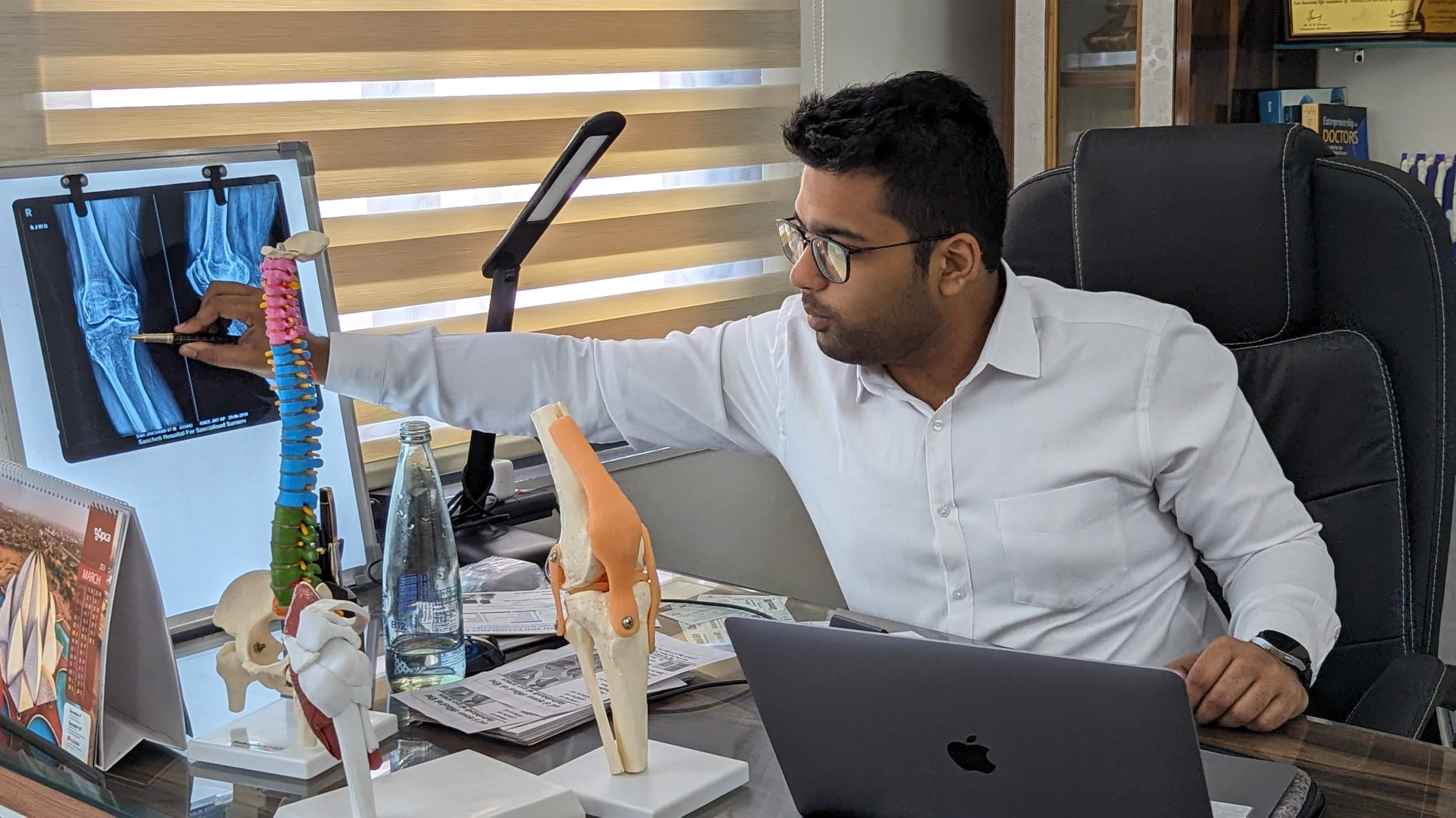 Dr. Siddharth Gupta reviewing a knee X-ray beside an anatomical knee model at his Faridabad clinic desk